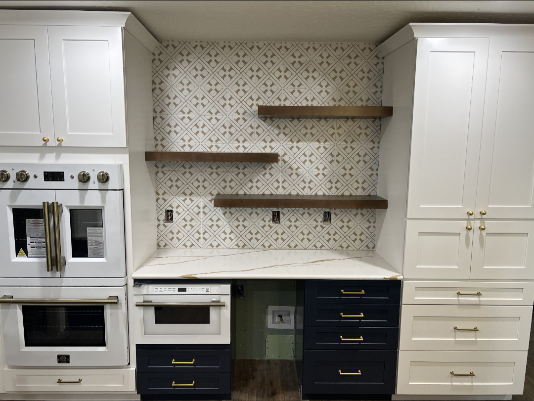 Remodeled two-tone kitchen with white shaker uppers, navy lower drawers with brass pulls, patterned tile backsplash, stained wood floating shelves and a French-door wall oven