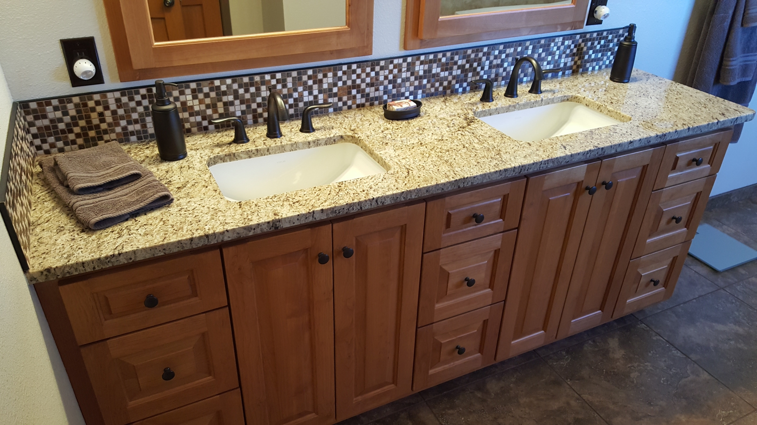 Double vanity with honey-oak cabinets, speckled granite counters, mosaic tile backsplash and bronze fixtures