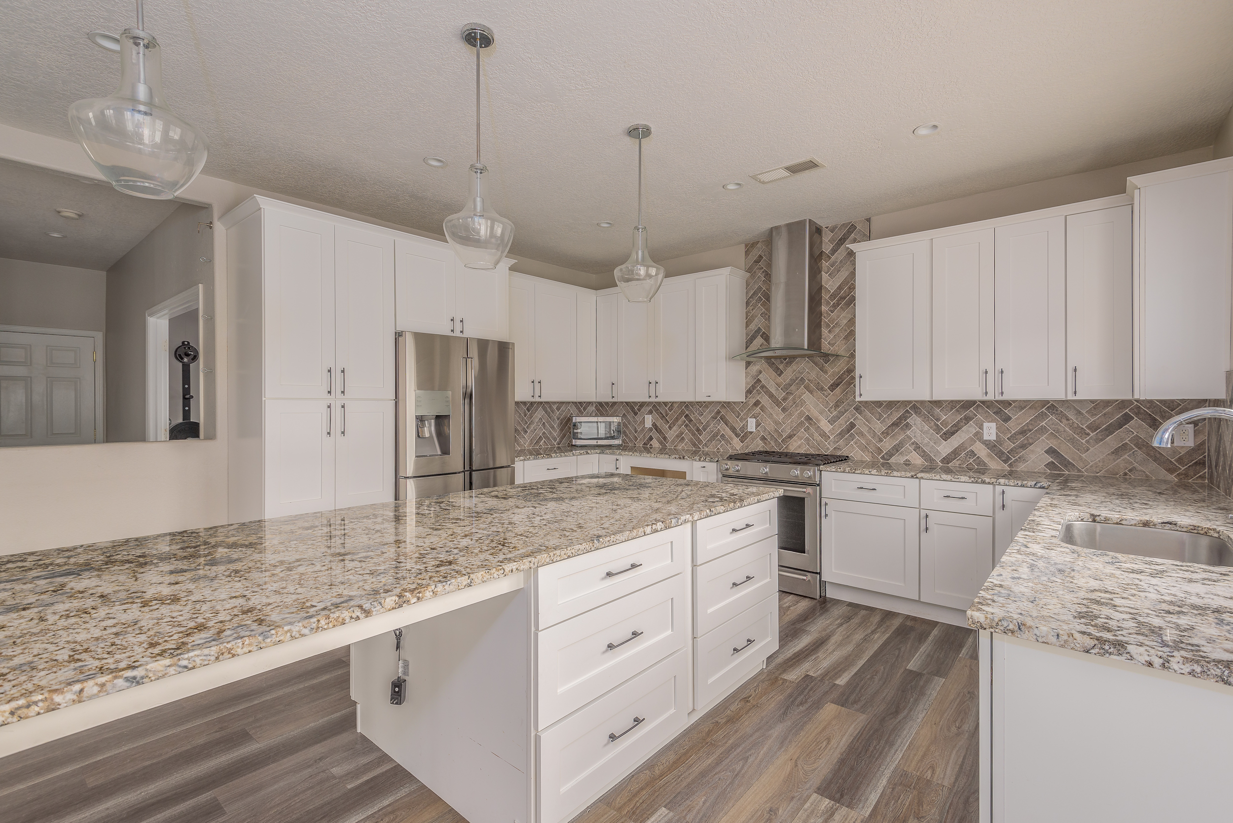 Bright white shaker kitchen with granite counters, herringbone tile backsplash, glass pendants and a large island